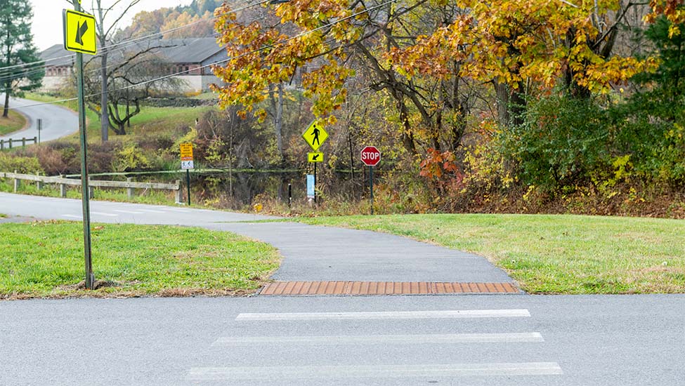 A pedestrian crosswalk leading to a winding path up a hill towards a barn in the distance.