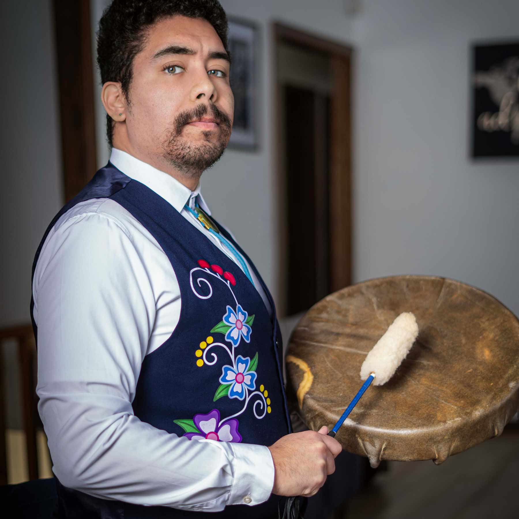 An individual stands holding a circular hand drum and a mallet with a soft white head. They wear a light-colored long-sleeve shirt under a navy vest decorated with colorful floral embroidery. The setting appears to be an indoor hallway.