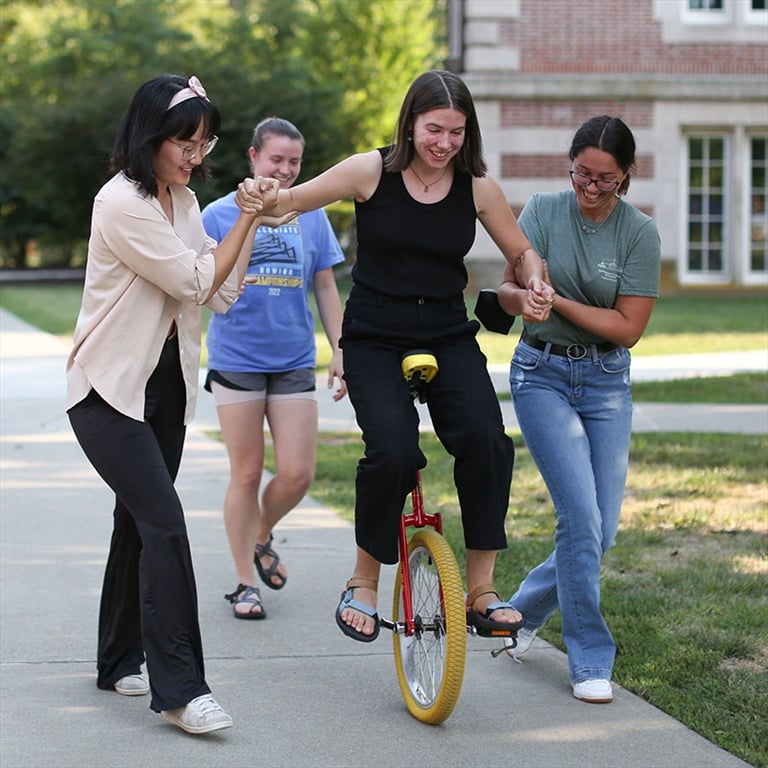Three students assist another student balanced on a unicycle on a path on Vassar's campus.