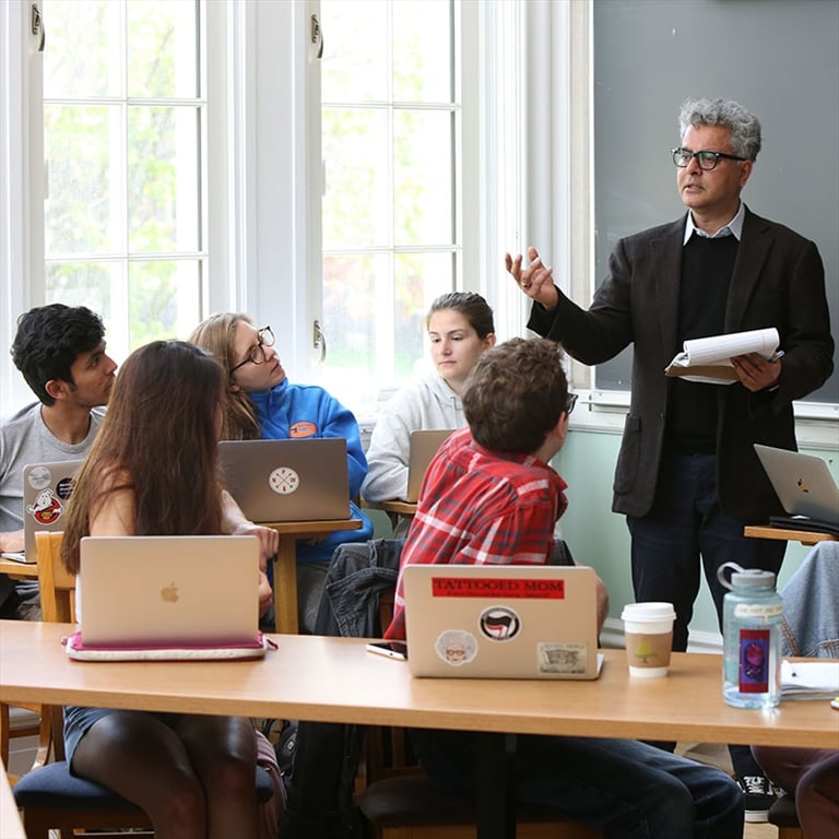 A professor with grey hair and glasses stands gesturing in front of a class of students seated at desks looking up at him.