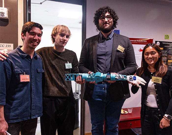 Four people standing indoors at a research poster session, smiling and holding a blue-and-white robotic arm prototype.