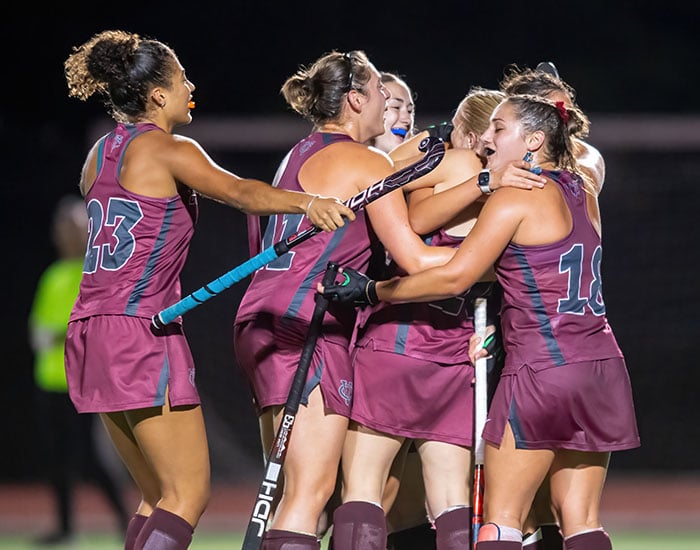 Field hockey players in maroon uniforms celebrating together on the field at night.