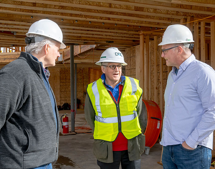 Three people wearing hard hats talk inside a building under construction, with one of them also wearing a reflective safety vest.