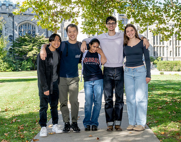 Five students stand arm in arm on a campus walkway under a leafy tree, with a Gothic-style academic building in the background.