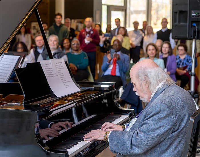 A pianist plays a grand piano during a performance, while an audience seated and standing in the background watches attentively.