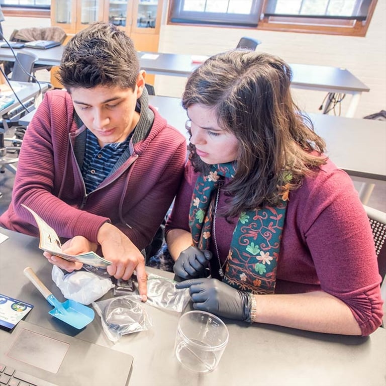 Two students confer while sitting at a table with a beaker, other items in plastic bags, and a laptop in front of them, in a classroom.