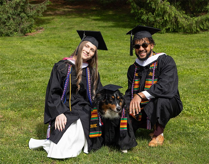 Two graduates in caps and gowns kneel on grass with a dog dressed in matching graduation attire sitting between them.