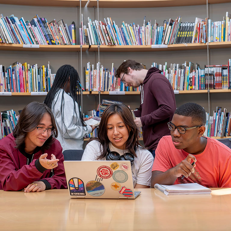 Three students sit at a desk with a laptop in front of them, talking, while two students stand in front of a bookcase behind them, looking at books.