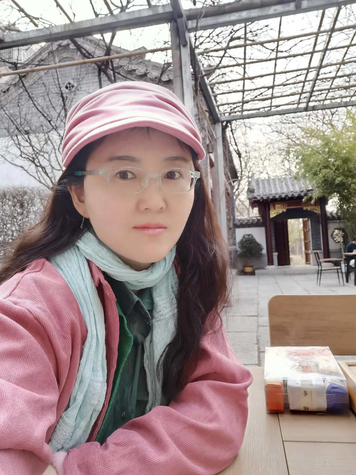 A figure is seated at a wooden table, wearing a pink cap and a light blue scarf over a layered outfit that includes a pink jacket and a green shirt. The figure is positioned slightly to the left of the frame, with arms crossed on the table. In the background, a wooden trellis can be seen above, partially obscured by bare branches, while hints of traditional architecture peek through, including a doorway framed in red. Soft, natural light illuminates the scene, creating a calm atmosphere.