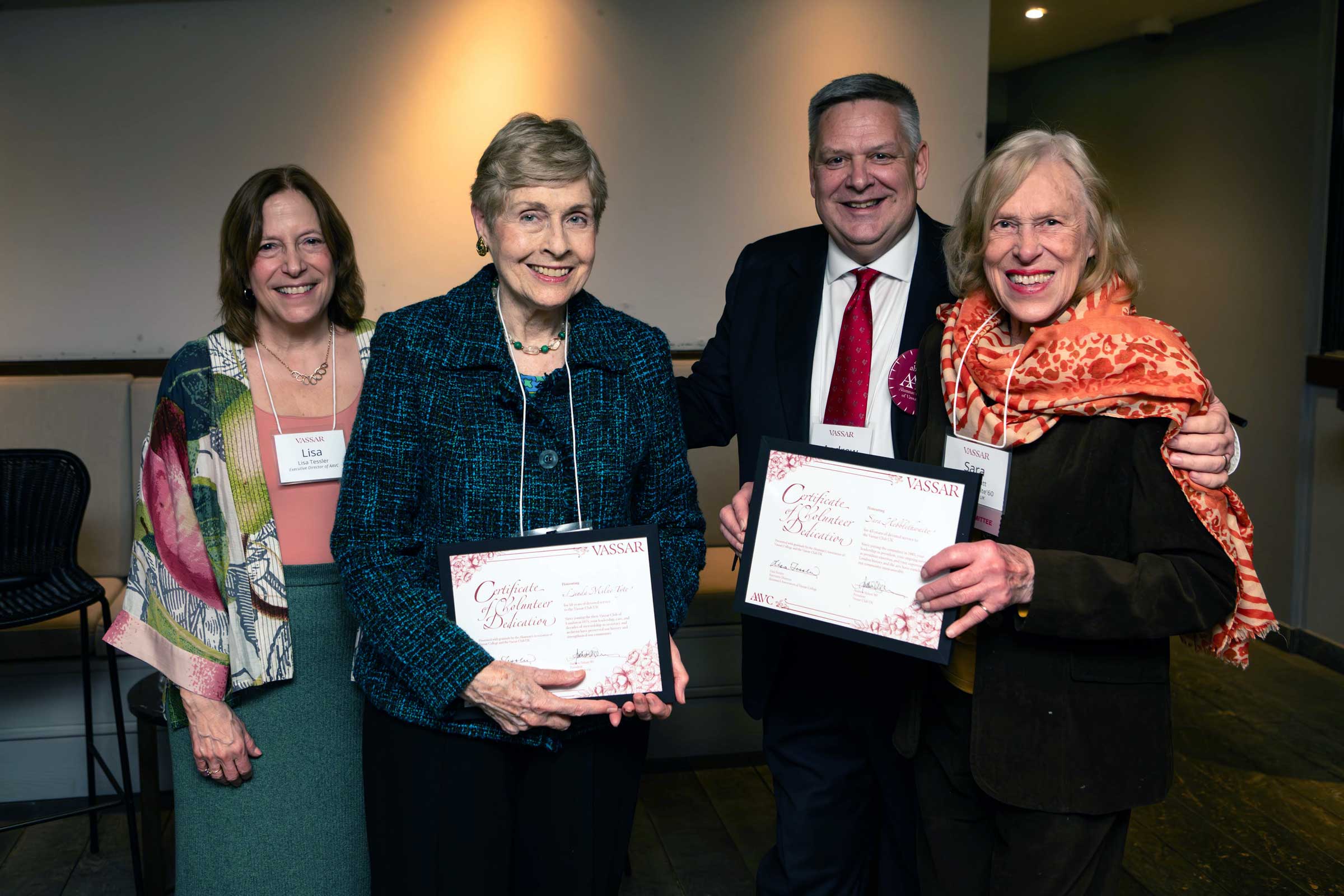 Four smiling people standing side by side while two in the center hold framed documents.