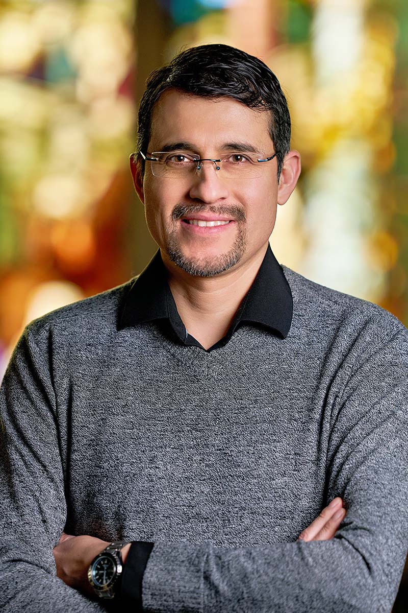 Juan Merlo with short dark hair, glasses, and a trimmed goatee, wearing a gray sweater over a black collared shirt, standing with arms crossed against a softly blurred, colorful background.