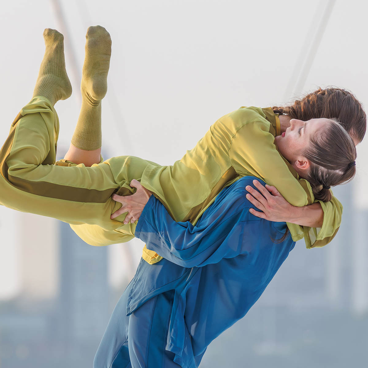 Two dancers perform a lift, with one dancer in a blue outfit holding another in a yellow outfit horizontally in their arms against a softly blurred outdoor background.