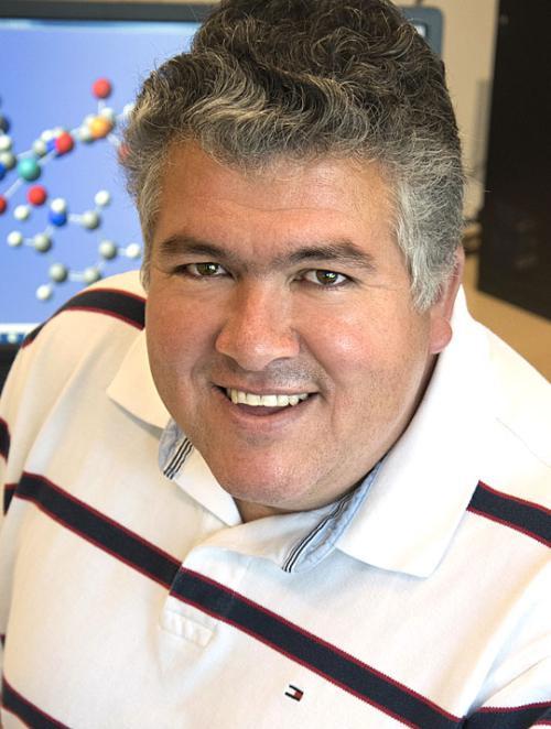 A headshot of a smiling person with short gray hair, wearing a white polo shirt, with a molecular model display visible in the background.