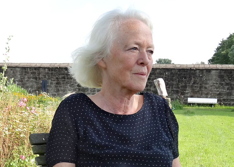 Portrait of Barbara Beisinghoff seated outdoors in a garden, wearing a dark patterned top, with flowers, grass, and a stone wall in the background.