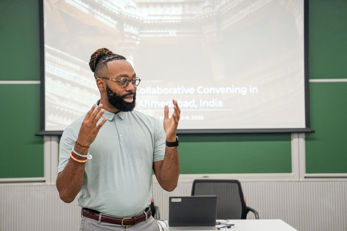 A person speaks, hands raised, in front of a projection screen in a conference room.