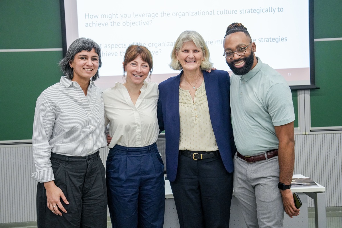 Four people stand in front of a conference room projector screen, smiling at the camera.