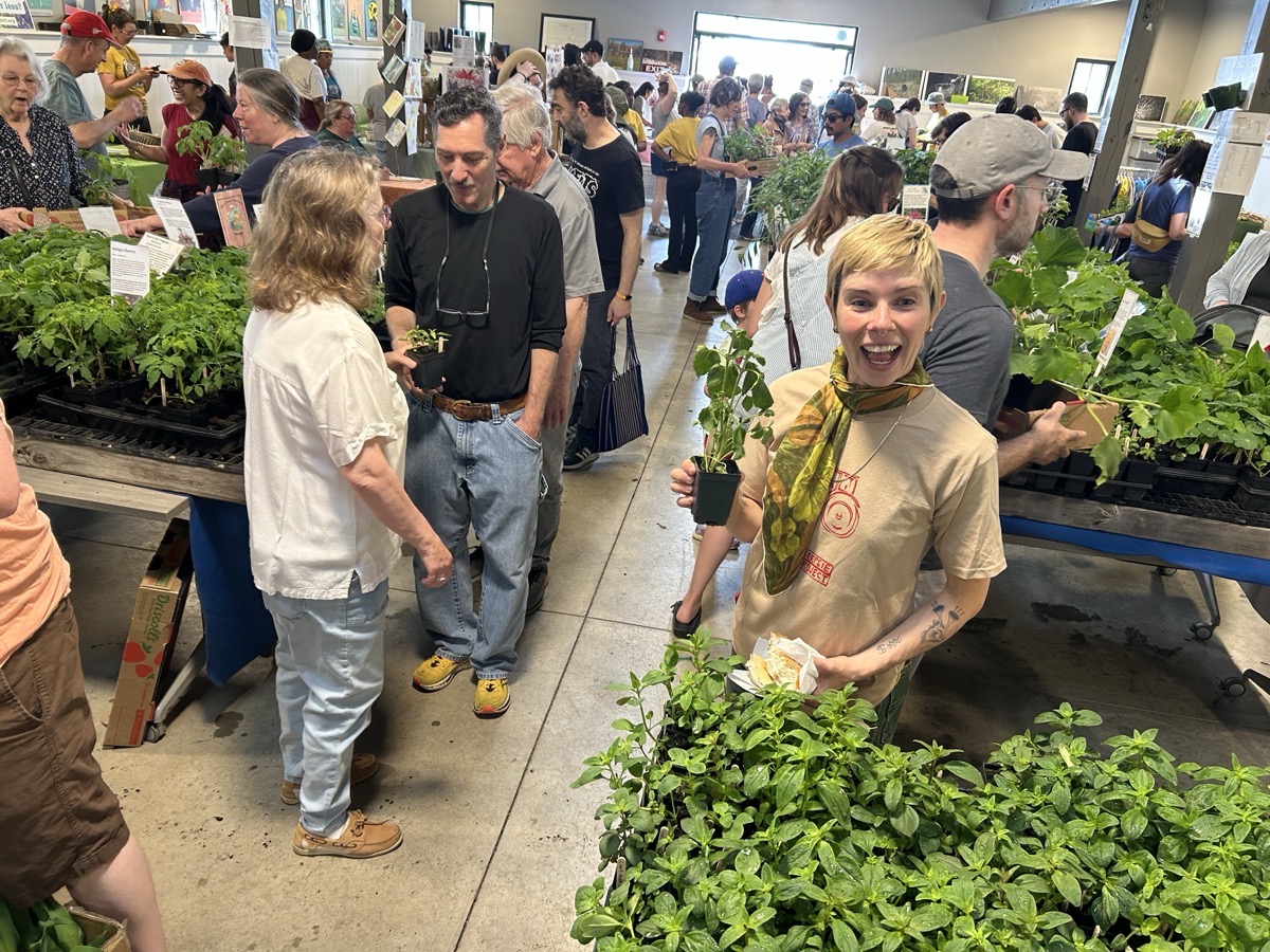 A vibrant indoor market scene filled with people engaged in browsing and purchasing plants. Numerous tables hold an array of green plants in pots, with a focus on herbs and vegetables, creating a lush atmosphere. In the foreground, a person holds a pot of greenery while smiling, dressed in a light-colored shirt and a patterned scarf. The lighting is bright and natural, illuminating the variety of colors from the plants and the casual interactions among the crowd.