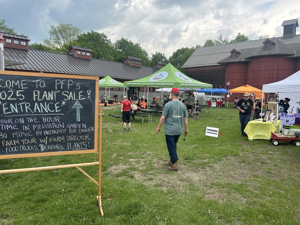 A chalkboard sign displays information about a plant sale, with text indicating activities and directions, positioned prominently in the foreground. In the background, various tents in green, orange, and blue are set up, with people seated at picnic tables and others walking among the vendors. The sky is partly cloudy, creating a soft, diffused light over the scene. Green grass underfoot contrasts with the dark barn structure behind, which features a sloped roof and multiple small windows.