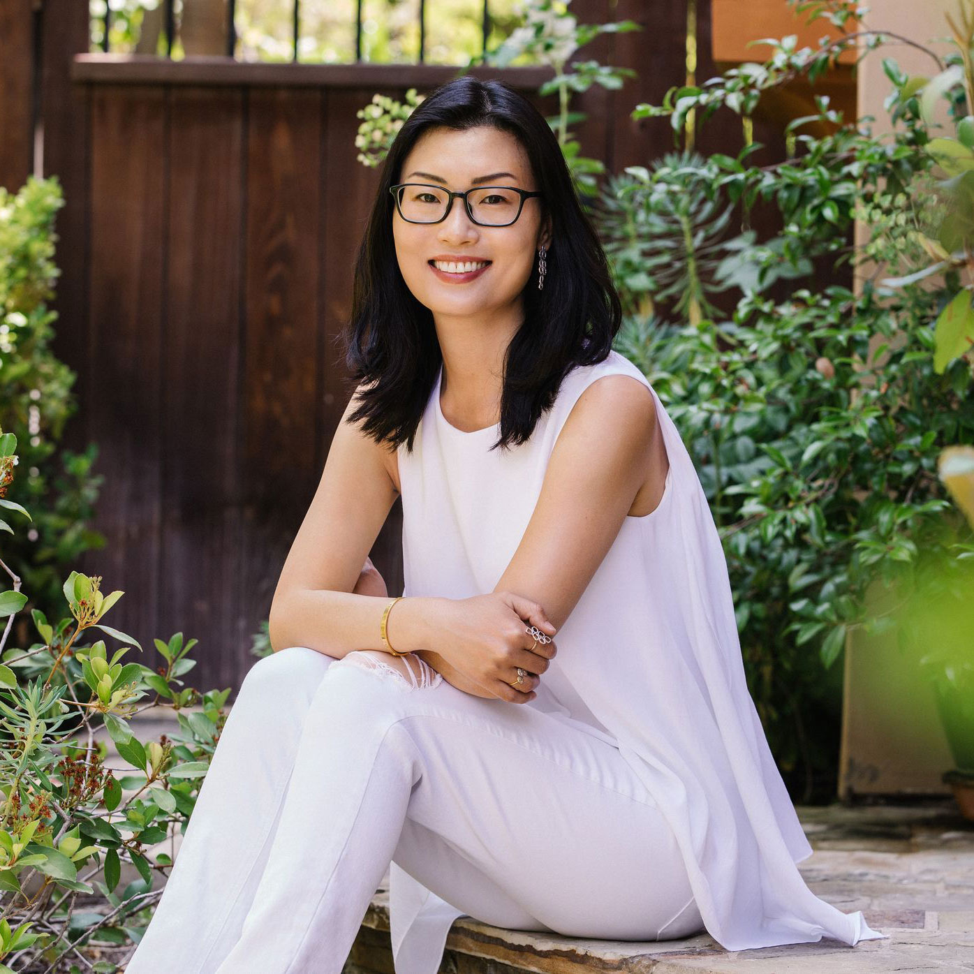 An individual with dark, shoulder-length hair and glasses smiles while sitting outdoors. They wear a white sleeveless top and white pants. Lush green plants and a wooden gate are in the background.