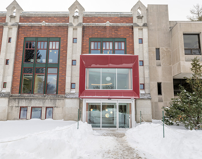 Three story brick building with tall windows and a modern red-framed entrance, surrounded by snow-covered ground.