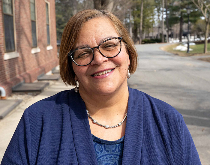 Linda Carter-Griffith ’84, a smiling person wearing glasses and a navy jacket, standing outdoors beside a brick building with trees in the background.