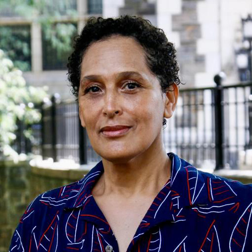 Head-and-shoulders portrait of a person outdoors, wearing a navy shirt with a red and white pattern, standing in front of a blurred background with a stone building and black iron fence.