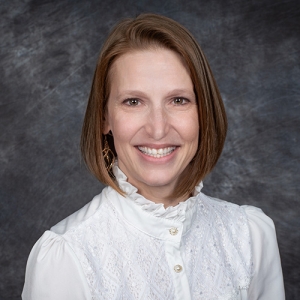 A headshot of a smiling person with shoulder-length brown hair, wearing a white lace blouse, against a mottled gray background.