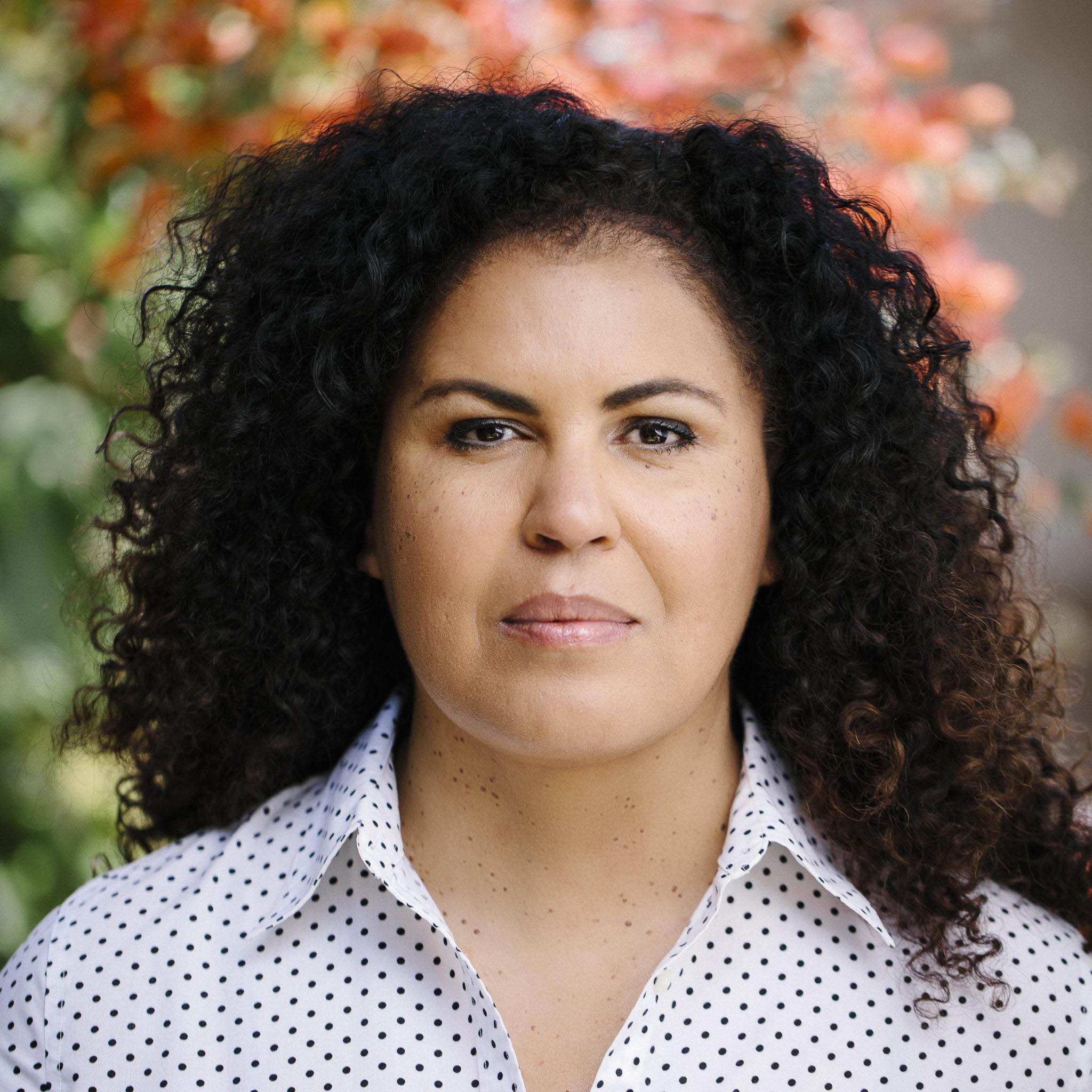 Head-and-shoulders portrait of Dr. Safiya U. Noble outdoors, wearing a white shirt with black polka dots, with curly dark hair and a softly blurred background of greenery and orange flowers. Photo credit: Stella Kallnina.