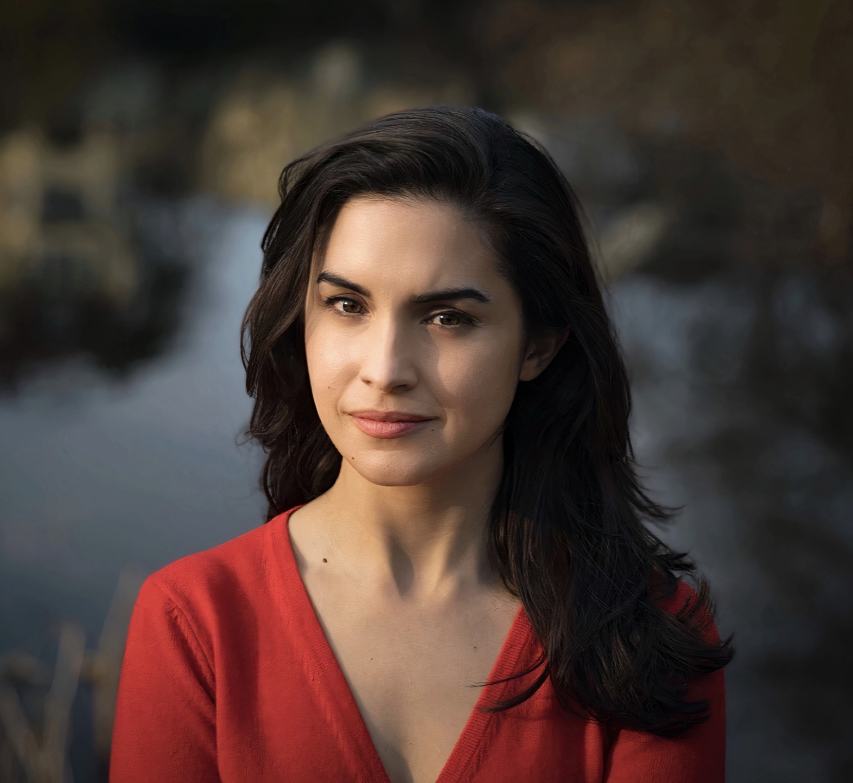 A photo of Merve Emre. They are dressed in a red top that contrasts with the blurred, dark background, which suggests a natural setting near water.