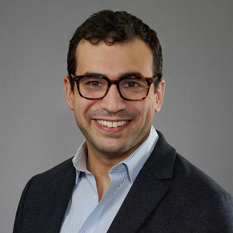 A headshot of a smiling person with short dark hair and tortoiseshell glasses, wearing a dark blazer over a blue striped shirt, against a neutral gray background.