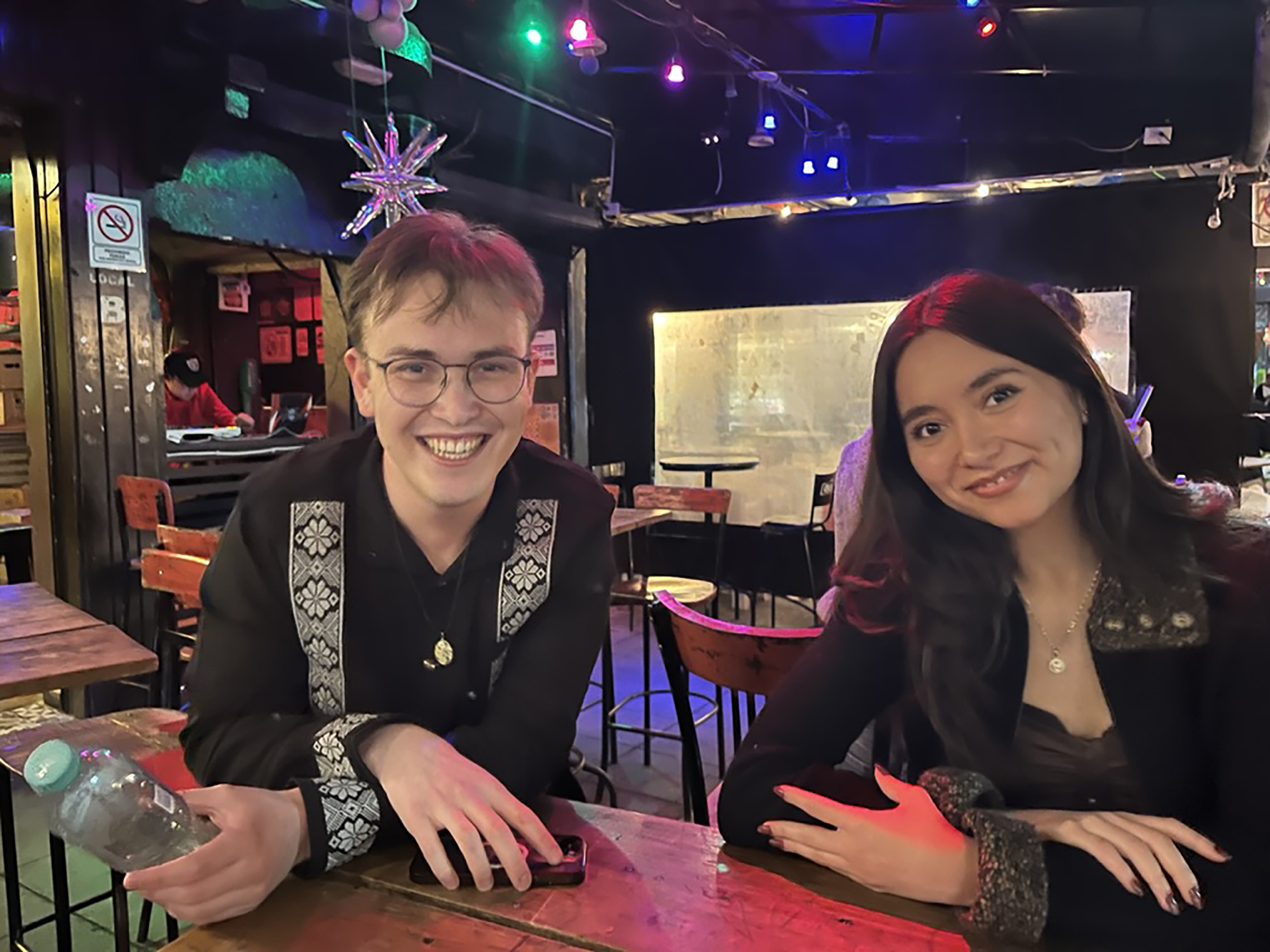 Two students smile at a restaurant table in a street market.