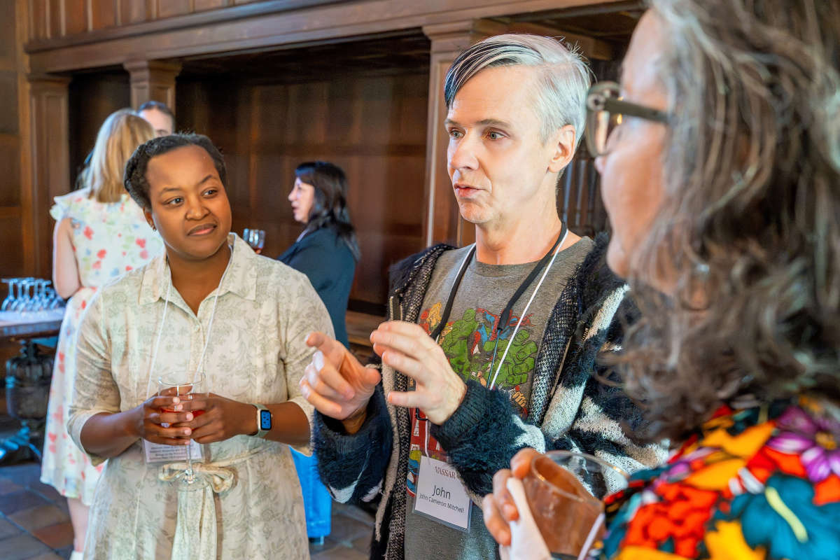 Several people wearing name tags talk and hold drinks during a social gathering in a room with wood-paneled walls.