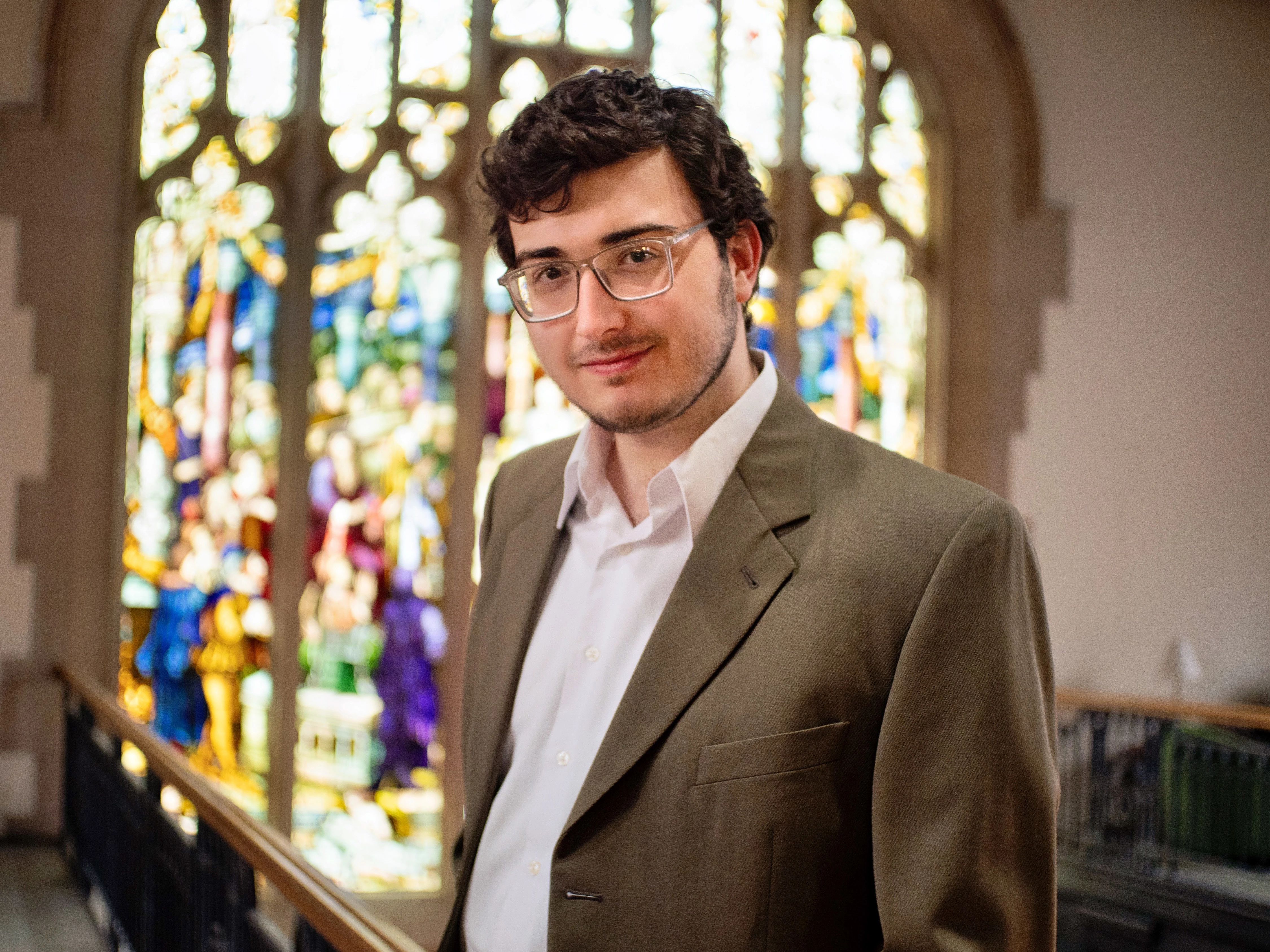 Bekir Hozdic ’26 stands in the Vassar library, with a stained glass window in the background. They are wearing glasses and a brown jacket.
