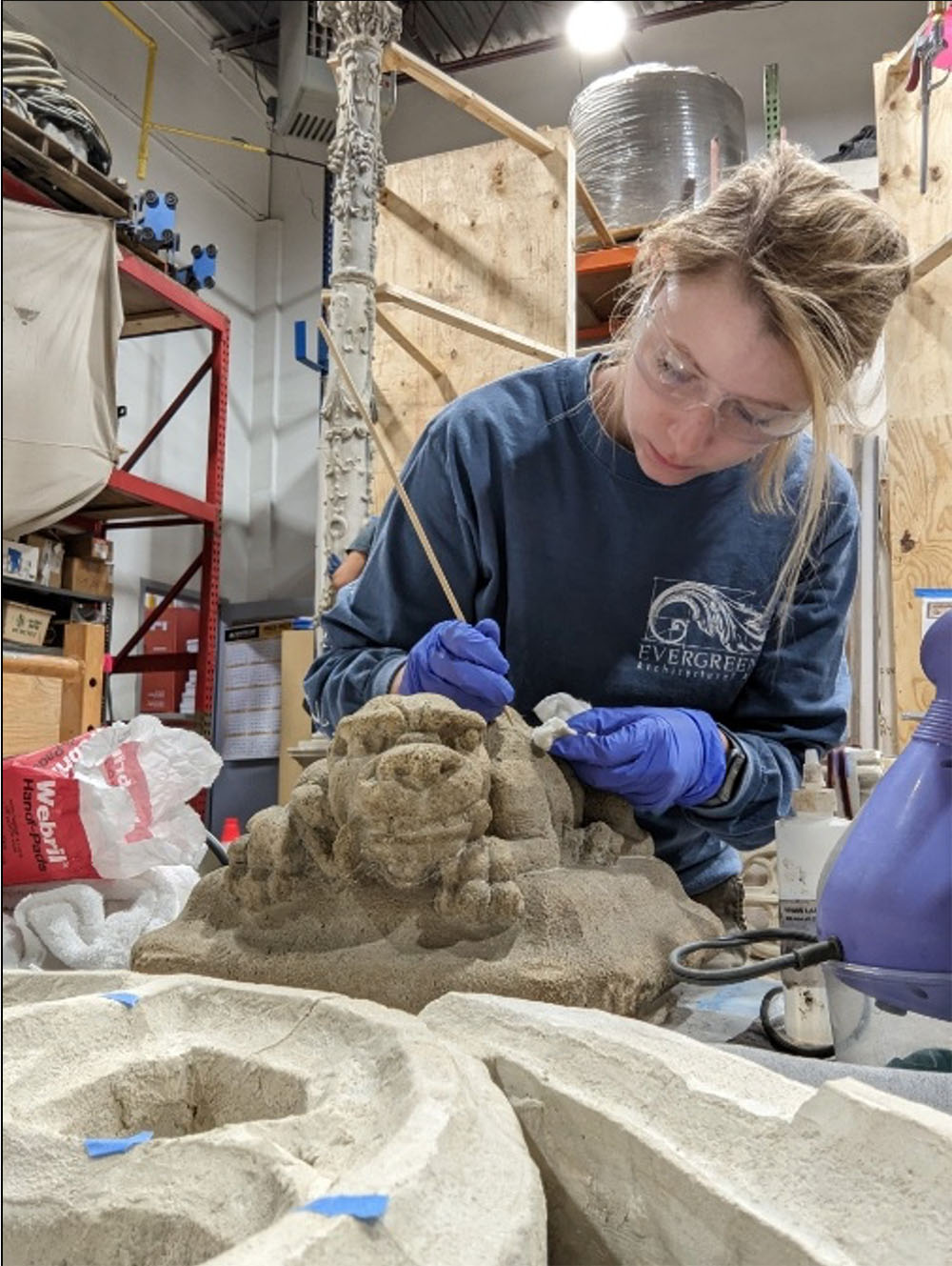 A conservator wearing gloves and safety glasses carefully cleans and restores a small stone sculpture using a fine tool in a workshop setting.