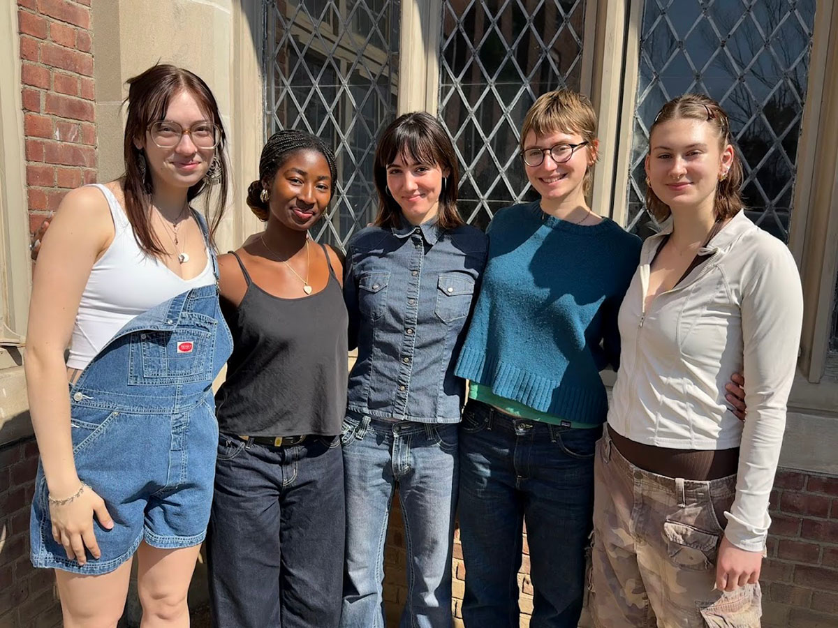 Five students stand together outdoors in front of a brick building with leaded glass windows, smiling, with their arms around each other.