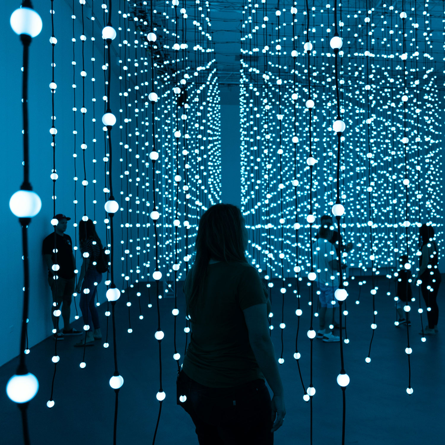 People in the dark walking among hundreds of strands of dimly lit light bulbs.