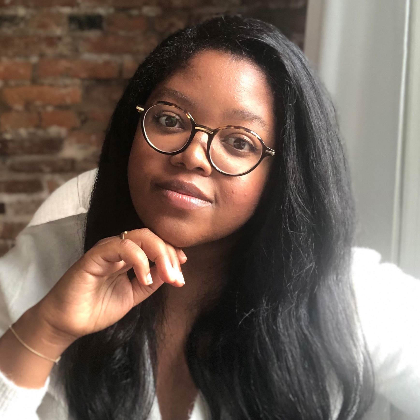 An individual with long dark hair and round tortoiseshell glasses looks at the camera with a hand resting beneath their chin. They wear a white top and a delicate gold bracelet and ring. An exposed brick wall and soft light frame the background.