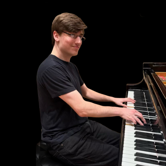 Person seated at a grand piano, playing the keys and looking toward the camera, wearing a black T-shirt against a dark background.