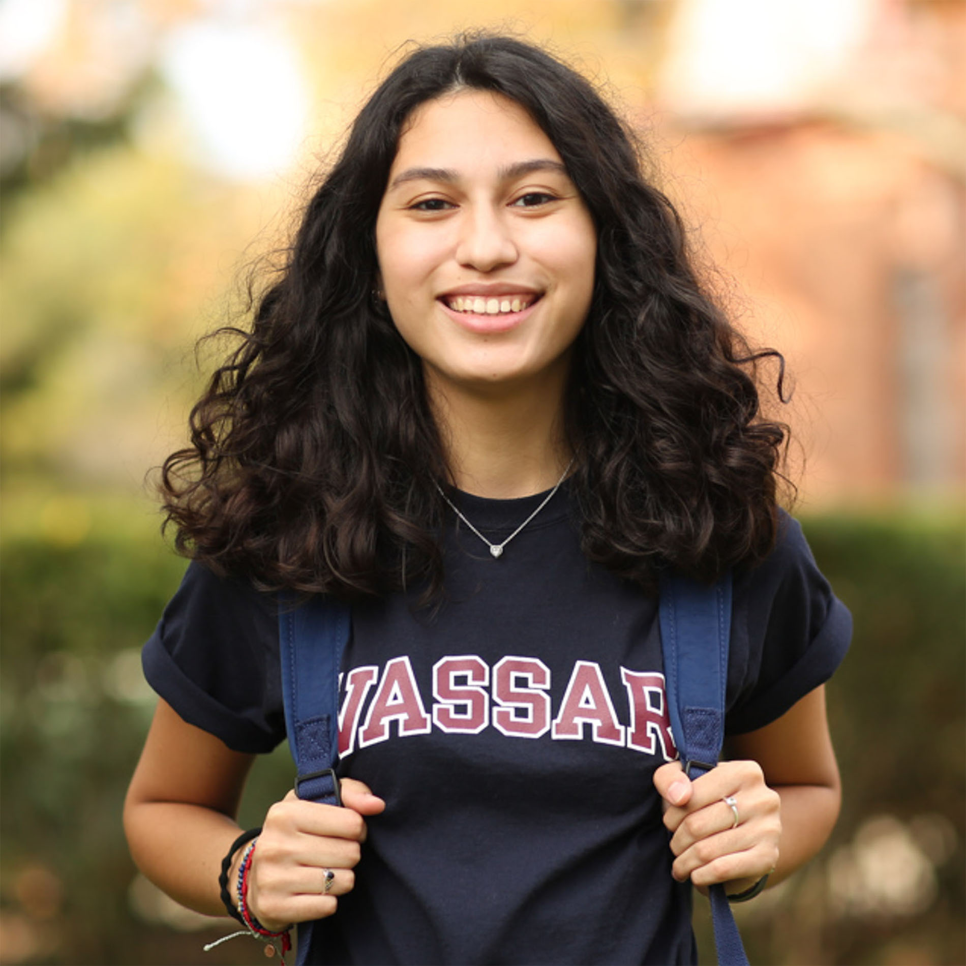 Person with long, dark, curly hair and a wide smile, wearing a "VASSAR" shirt and a backpack, outdoors.