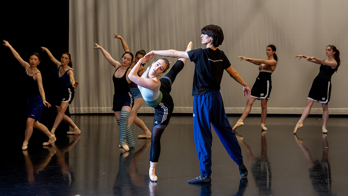 Dancers rehearsing in a studio, with one dancer extending in a supported pose while others practice movements in the background.