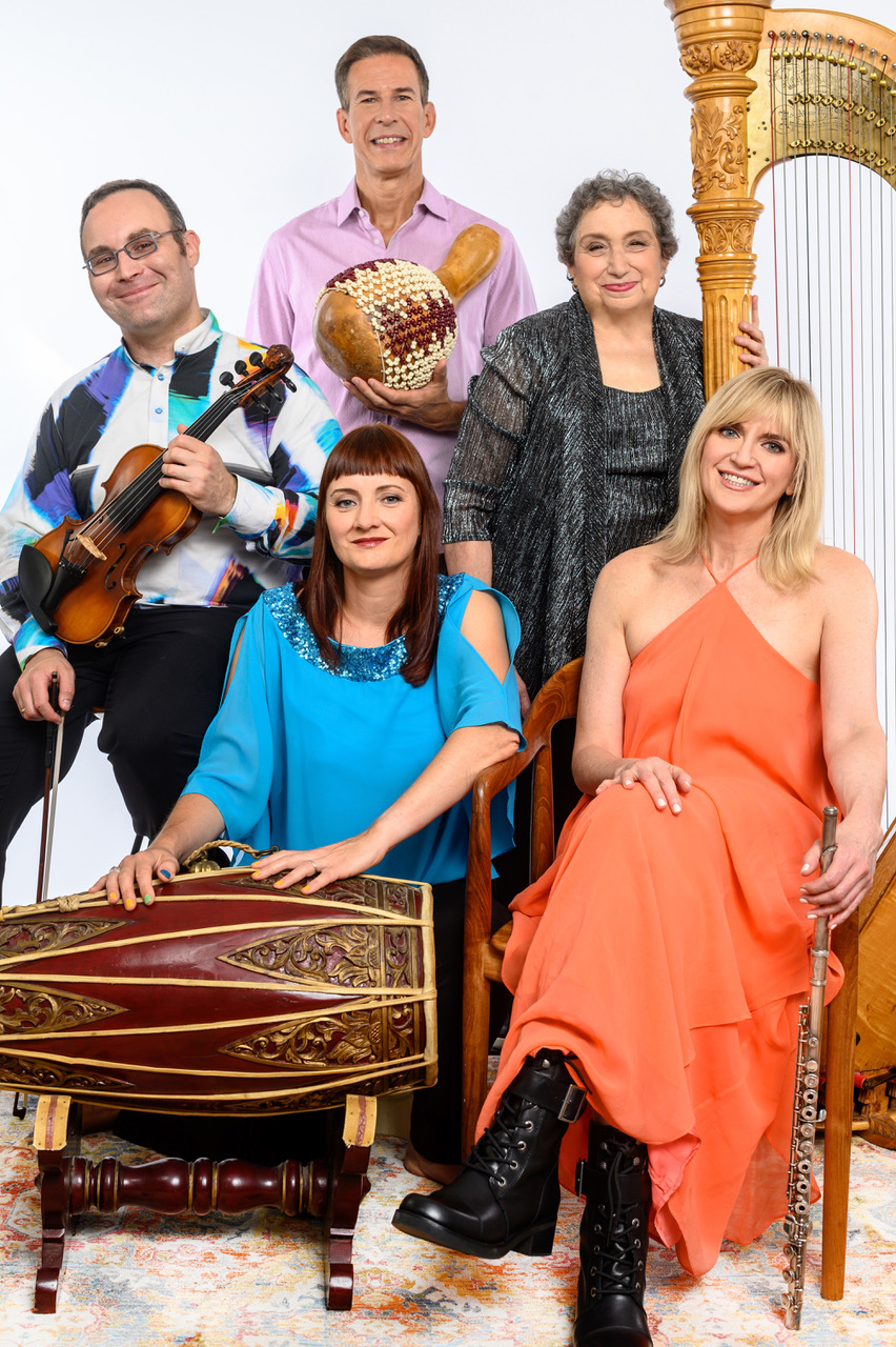 Group portrait of five musicians posing with their instruments against a light gray background. One person holds a violin, another stands holding a beaded gourd percussion instrument, and a large harp is visible at the right side of the image. Two seated performers wear bright blue and coral outfits, while the standing performers wear patterned and metallic attire. The group smiles toward the camera in a formal promotional-style photo.