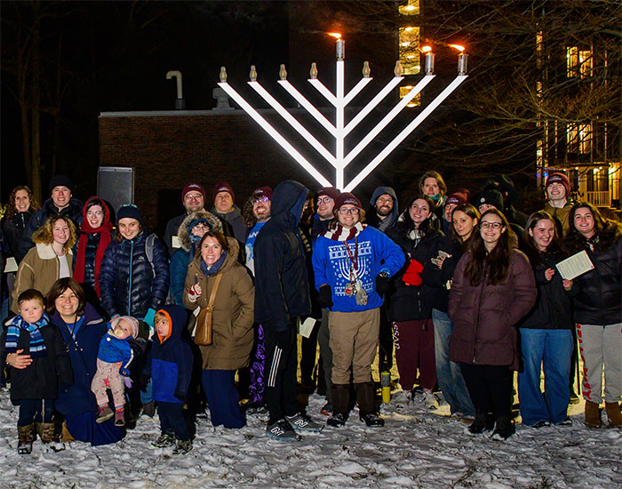 Group of people gathered outdoors in the snow at night around a large lit menorah, posing together for a photo.