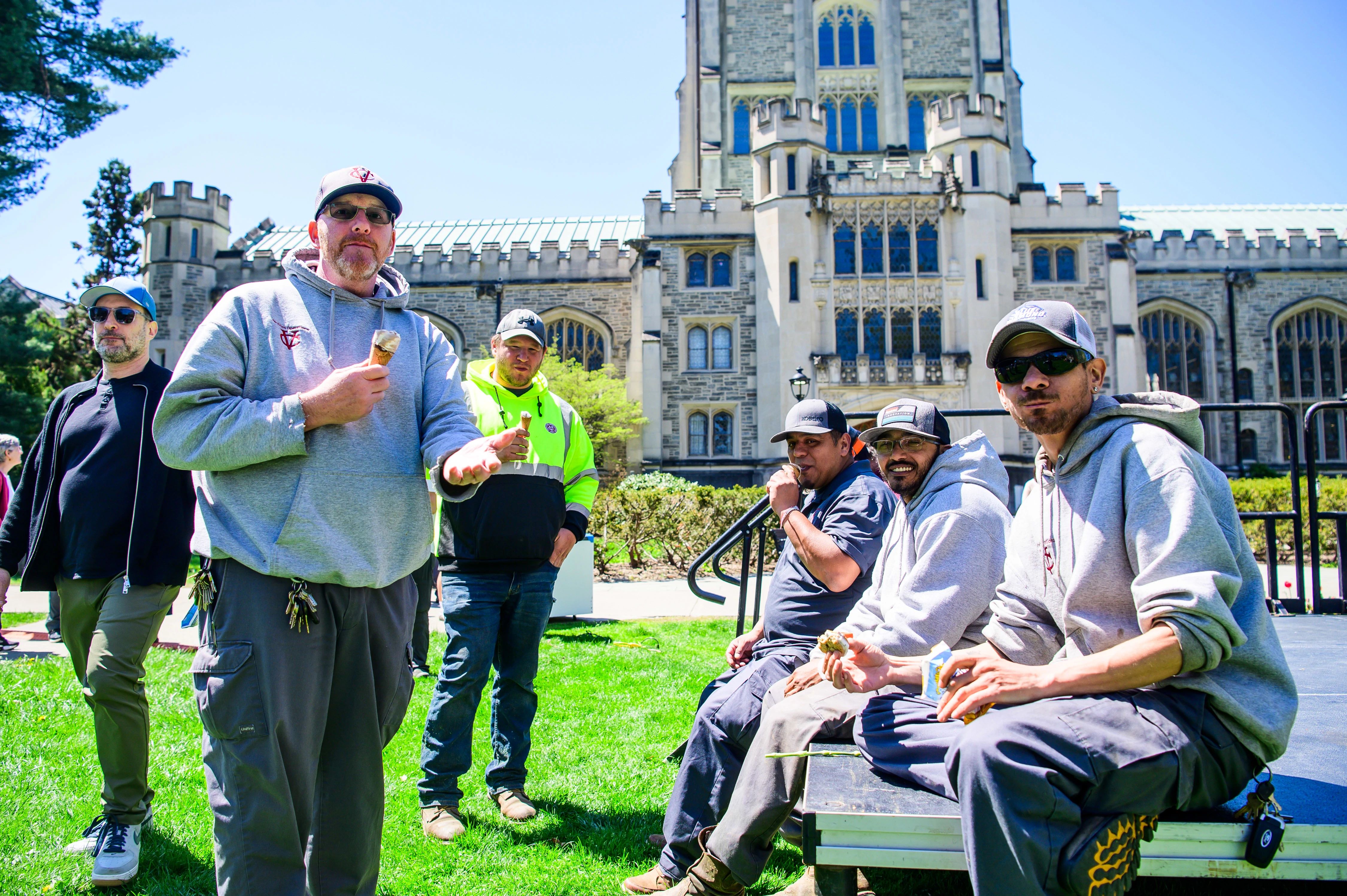 Several members of the grounds crew stand and and sit on the librrry lawn, eating ice cream. 