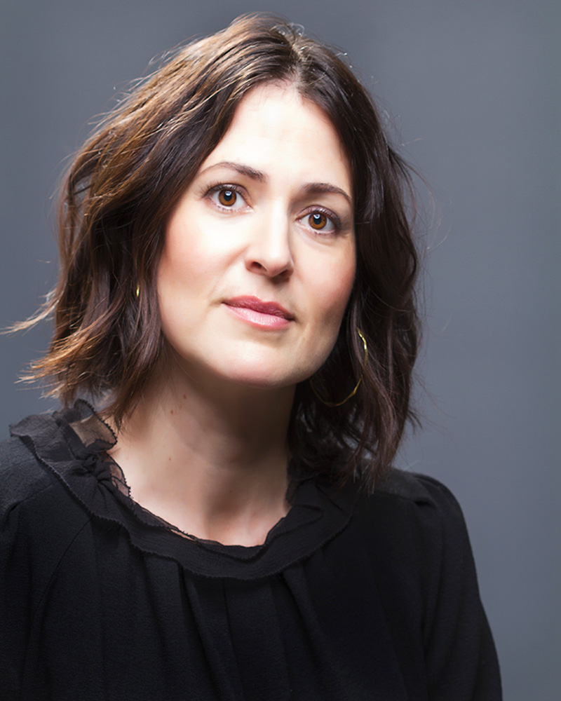 Alexandra Shiva with shoulder-length, wavy dark hair and wearing a black blouse with a delicate ruffled neckline and gold hoop earrings, against a neutral grey backdrop.