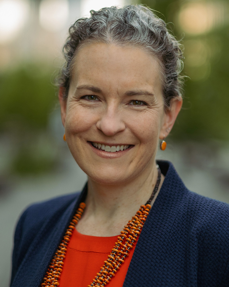 Anne Green, with curly salt-and-pepper hair, smiling warmly, wearing an orange top under a navy blue blazer, paired with a vibrant orange beaded necklace and matching earrings. The background is a soft-focus outdoor setting.