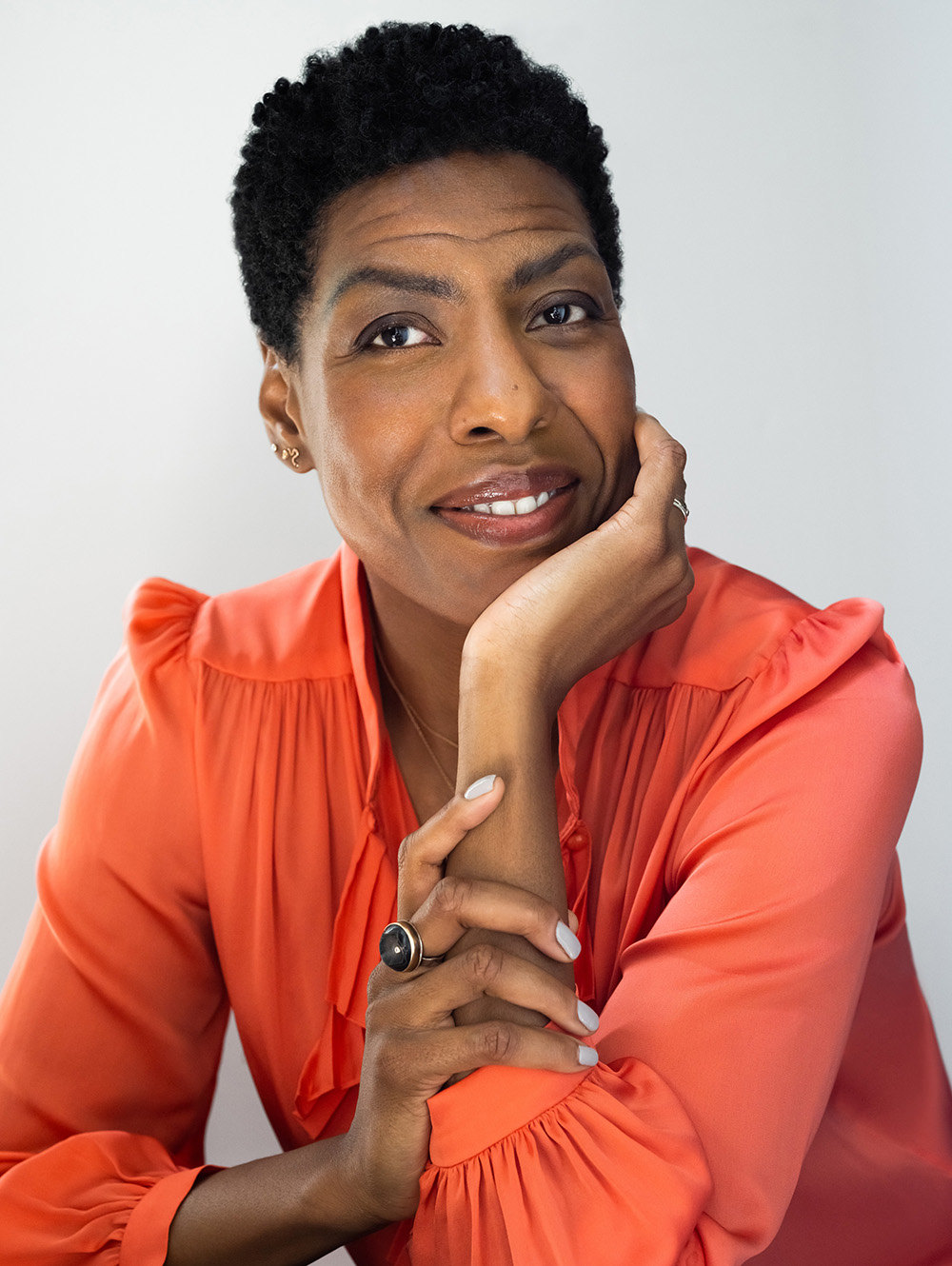 Ayana Mathis with short natural hair wearing a coral blouse, resting their chin on one hand and smiling against a light gray background.