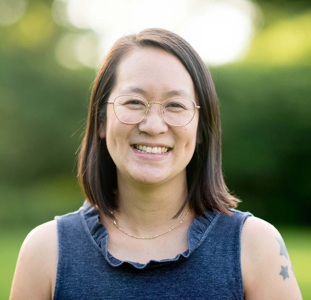 Delia Cheung Hom smiling warmly, wearing round glasses, a dark blue top with a ruffled neckline, and has a star tattoo visible on her left shoulder. The background is a soft-focus, bright green outdoor setting.