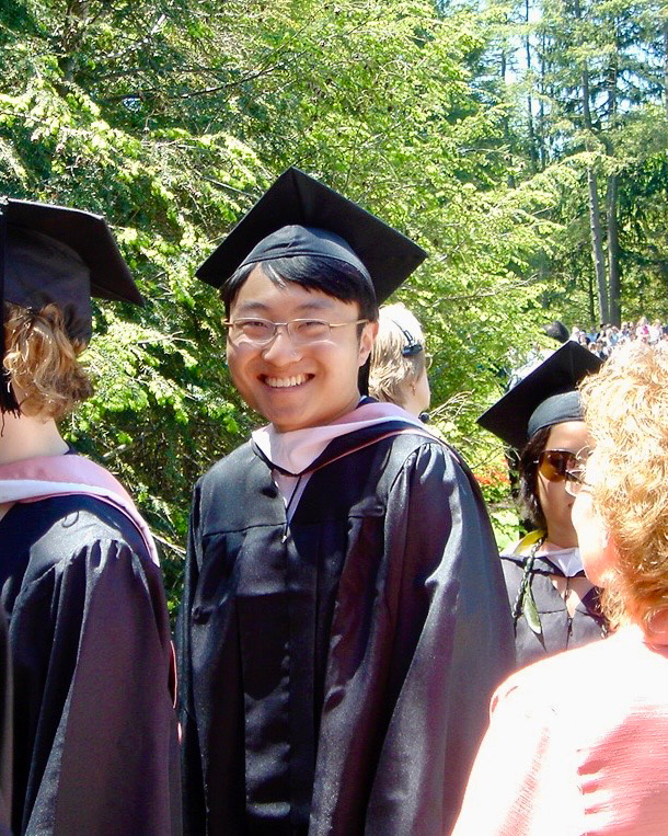 Eric Liu smiling in his graduation cap, a light pink academic hood, and gown during an outdoor commencement ceremony with green trees and other graduates in the background.
