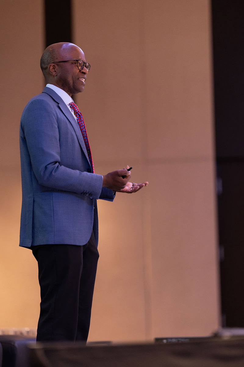 Kimathi Marangu giving a presentation on stage, wearing a grey blazer and red patterned tie while gesturing with his hands against a neutral, warm-toned paneled background.