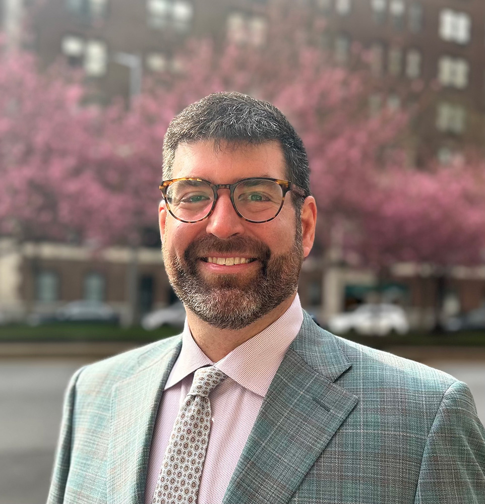 Winston Clement featuring a short beard and glasses, and dressed in a light grey plaid blazer, a pink patterned dress shirt, and a grey tie. The background is a soft-focus urban setting with blooming cherry blossom trees and brick buildings.